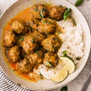 A bowl of rice topped with thai curry meatballs in a rich, orange-colored sauce, garnished with fresh basil leaves and a lemon wedge, served on a decorative plate with a fork on the side.