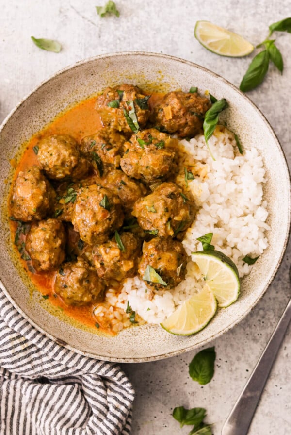 A bowl of rice topped with thai curry meatballs in a rich, orange-colored sauce, garnished with fresh basil leaves and a lemon wedge, served on a decorative plate with a fork on the side.
