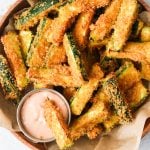 an overhead shot of air fryer zucchini fries in a brown bowl with one dipping into a sauce.