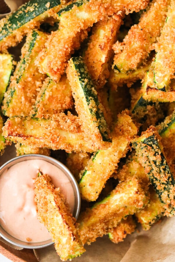an overhead shot of air fryer zucchini fries in a brown bowl with one dipping into a sauce.