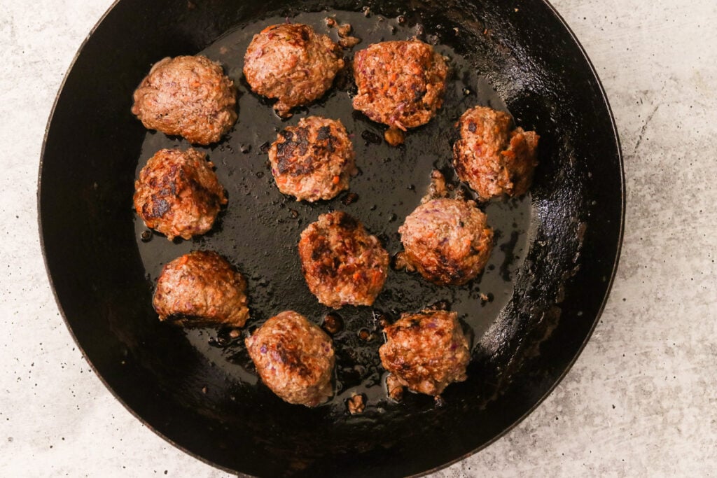 Pan frying meatballs in a black skillet until browned on all sides.