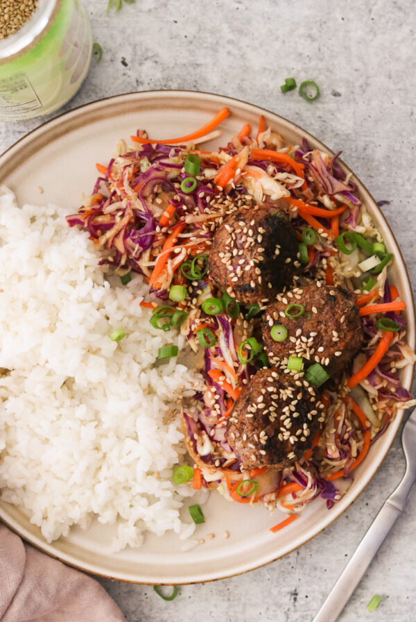 Egg roll meatballs served on a gray plate with white rice and sesame slaw, garnished with green onions and toasted sesame seeds.