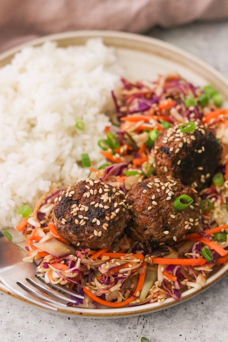 Egg roll meatballs served on a gray plate with white rice and sesame slaw, garnished with green onions and toasted sesame seeds.