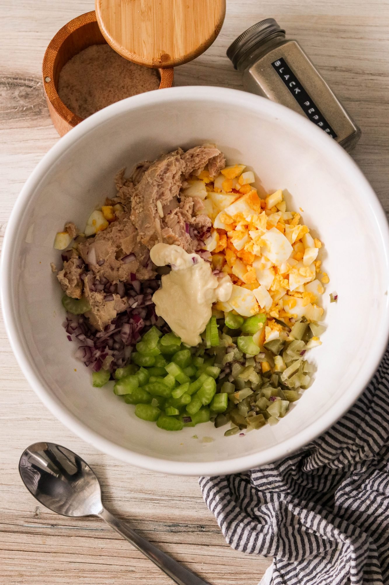 Overhead shot of ingredients for tuna egg salad in a white mixing bowl including tuna, hard-boiled eggs, celery, red onion, and pickles.
