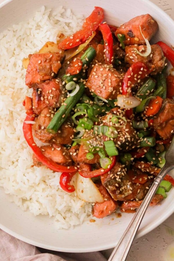 an overhead view of a white plate filled with white rice and salmon stir fry with a silver fork resting on the side of the plate