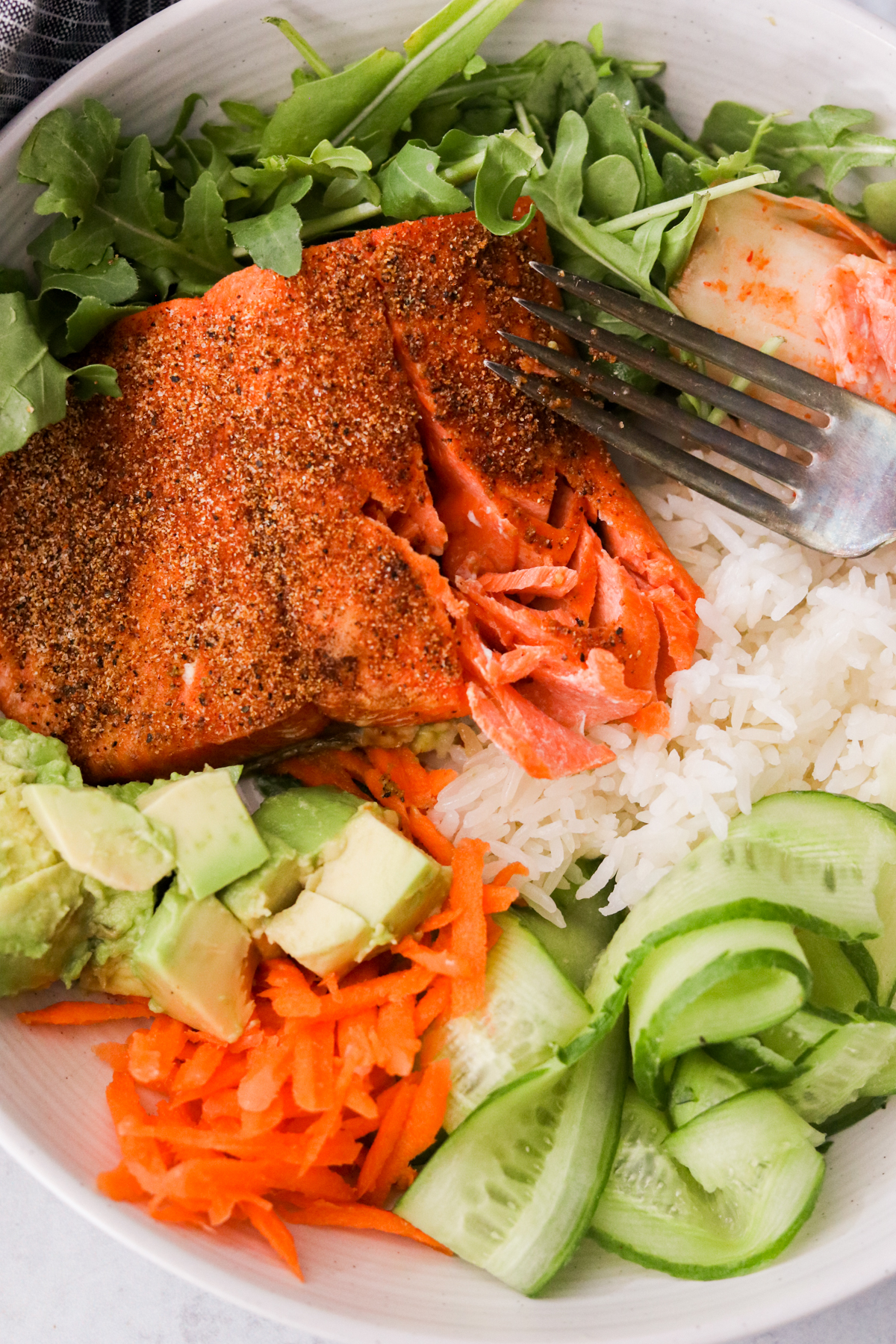 a close up of a cooked salmon fillet on a bed of white rice, with avocado, kimchi, and cucumbers