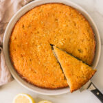 gluten-free lemon drizzle cake in a round cake pan one triangle slice cut and resting on a knife