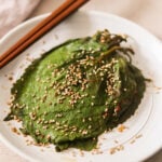 a plate of picked perilla leaves on a white plate topped with sesame seeds and chopsticks resting on the top..