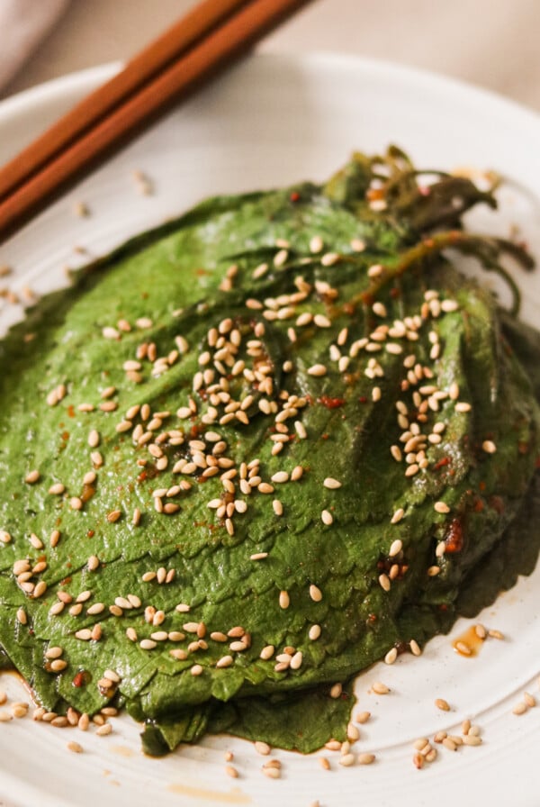 a plate of picked perilla leaves on a white plate topped with sesame seeds and chopsticks resting on the top..