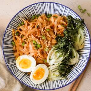 chili garlic noodles in a blue bowl and topped with soft boiled eggs and roasted bok choy