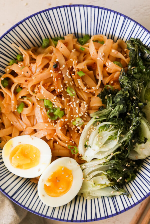 chili garlic noodles in a blue bowl and topped with soft boiled eggs and roasted bok choy