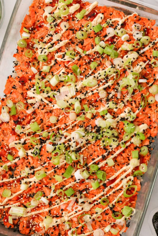 a salmon sushi bake in a casserole dish topped with mayo, sesame seeds, and green onions.