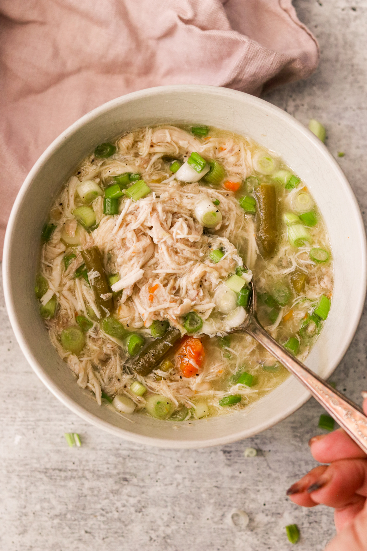 a hand dipping a spoon into a bowl of soup filled with shredded chicken and veggies