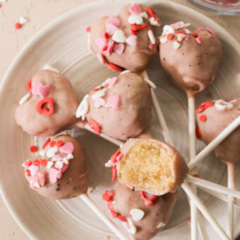 7 Valentine's cake pops on a white plate and topped with heart shaped sprinkles and a bite taken out of one to show the middle