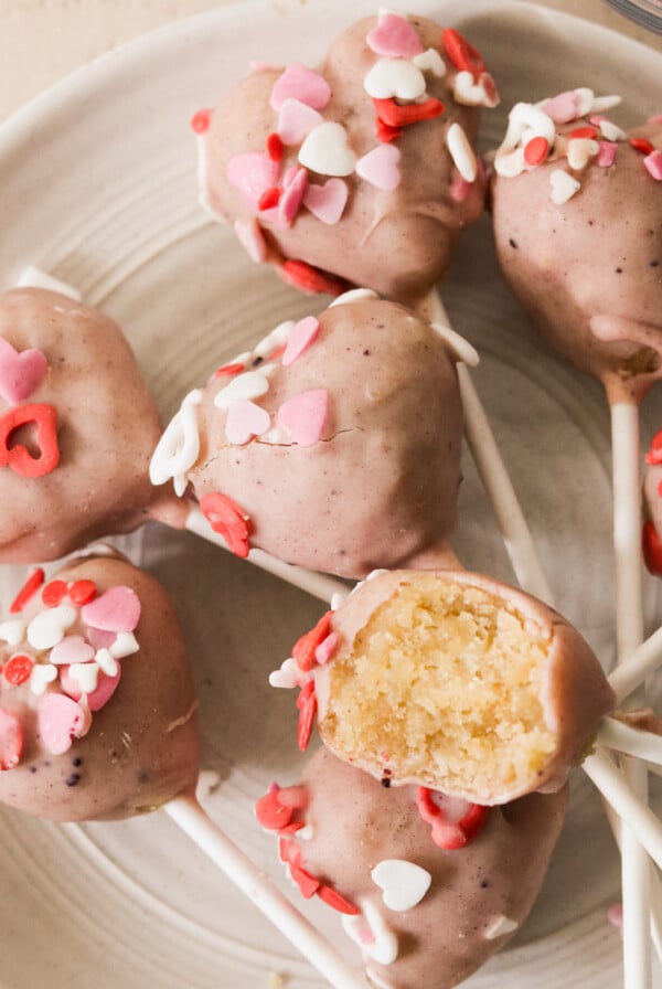 7 Valentine's cake pops on a white plate and topped with heart shaped sprinkles and a bite taken out of one to show the middle