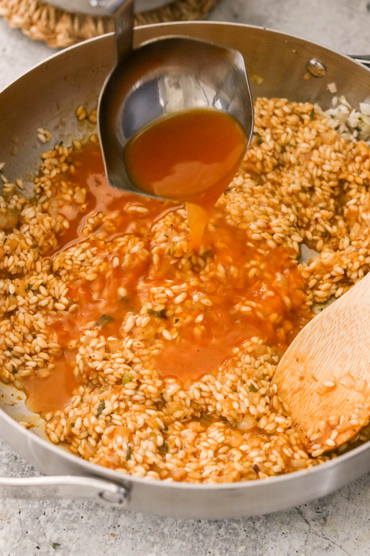 A ladle pouring broth into a pan of arborio rice and herbs.