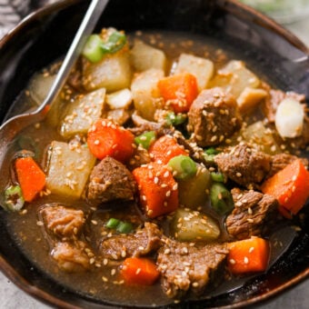 Close-up of a hearty Asian beef stew with slow-cooked beef, carrots, and daikon in a savory broth, topped with sesame seeds and fresh green onions.