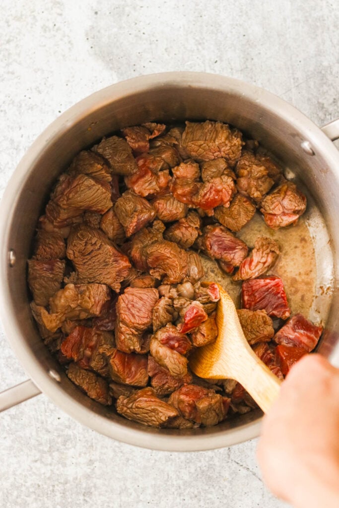 Cooking beef chunks in a pot with a wooden spoon, browning the meat for Asian beef stew.