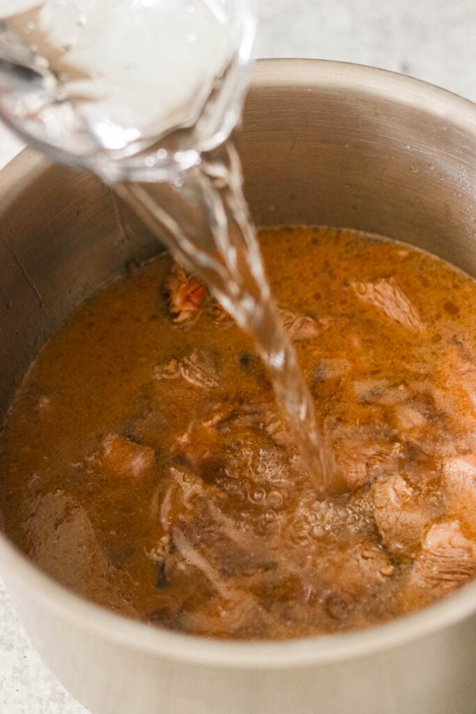 Pouring water into a simmering pot of Asian beef stew to create a rich and flavorful broth.