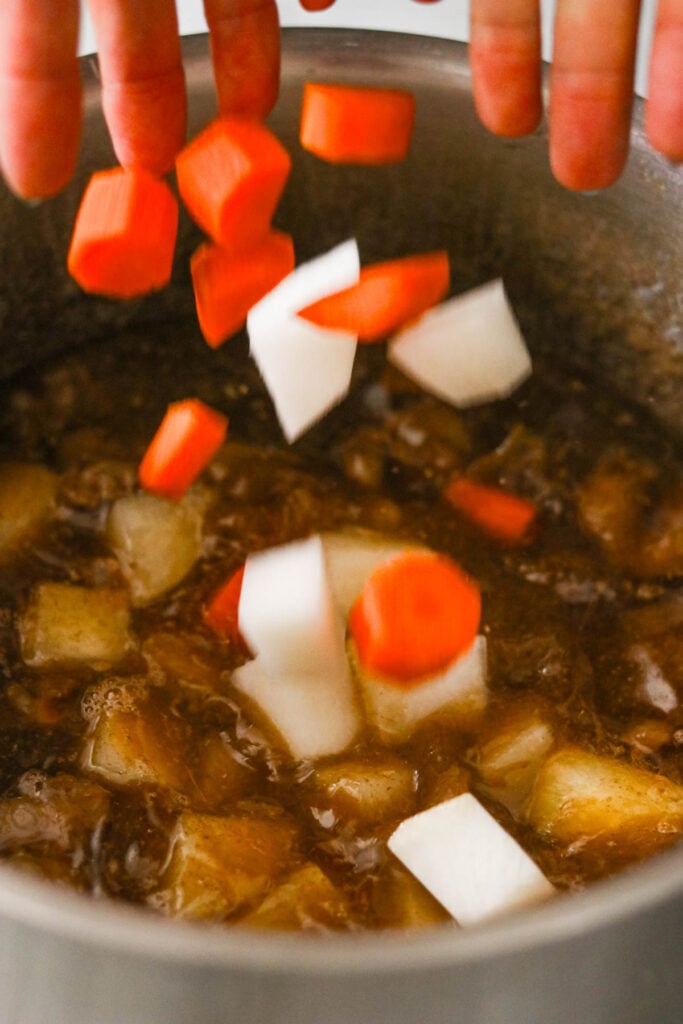 Hands tossing chopped carrots and daikon radish into a pot of simmering beef stew.