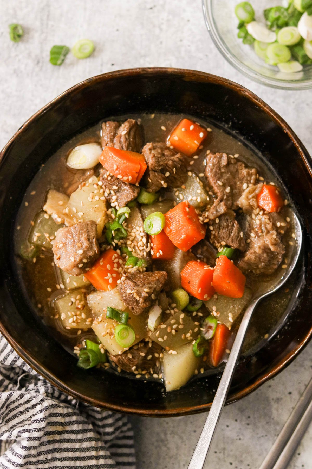 A bowl of Asian beef stew with tender beef chunks, carrots, daikon radish, and sesame seeds, garnished with green onions, served in a dark ceramic bowl.