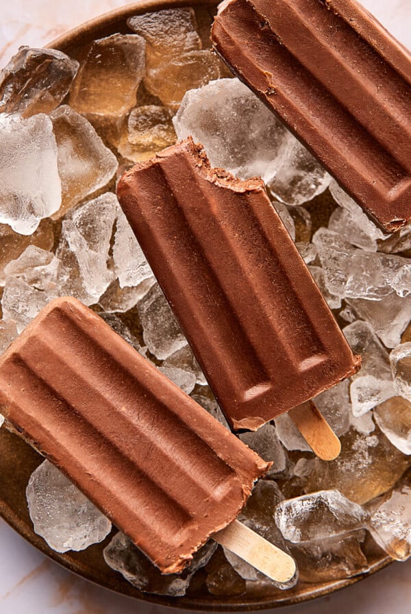 Three chocolate fudgesicles, with one having a bite taken out, are placed on a plate filled with ice cubes. The background features a light marble surface.