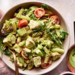 A bowl of green goddess pasta salad with cherry tomatoes, avocado, arugula, and a creamy green dressing. A glass of water with lemon slices and a small bowl of dressing with a wooden spoon are nearby. Lemon slices and fresh greens are scattered around.