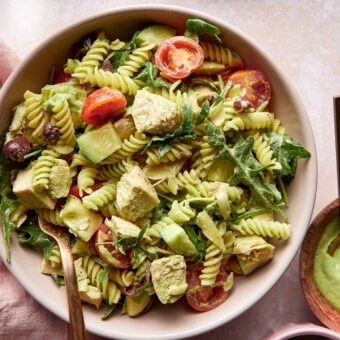 A bowl of green goddess pasta salad with cherry tomatoes, avocado, arugula, and a creamy green dressing. A glass of water with lemon slices and a small bowl of dressing with a wooden spoon are nearby. Lemon slices and fresh greens are scattered around.