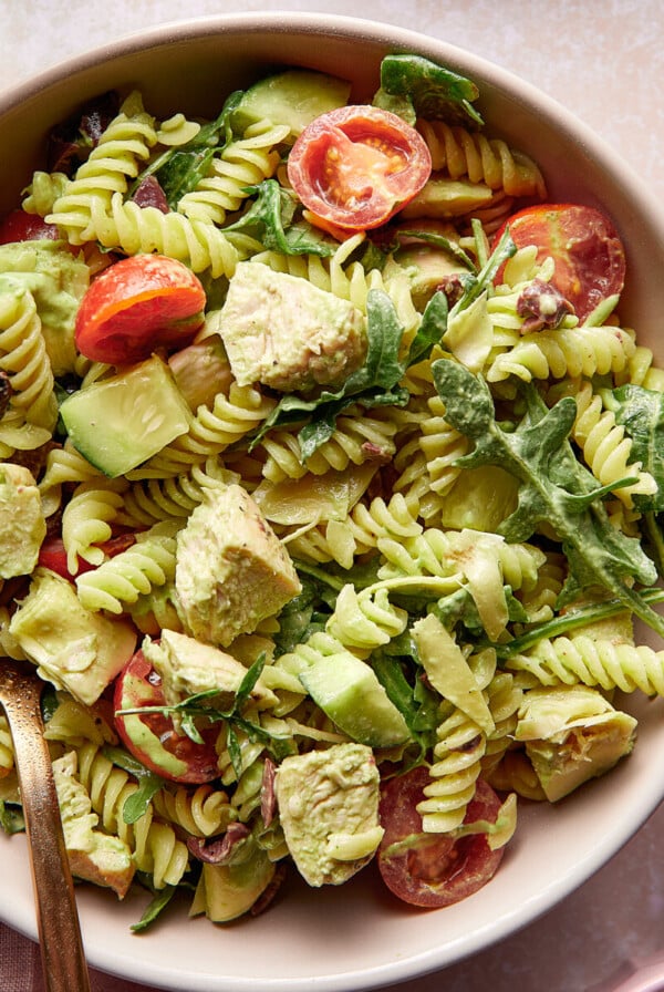 A bowl of green goddess pasta salad with cherry tomatoes, avocado, arugula, and a creamy green dressing. A glass of water with lemon slices and a small bowl of dressing with a wooden spoon are nearby. Lemon slices and fresh greens are scattered around.