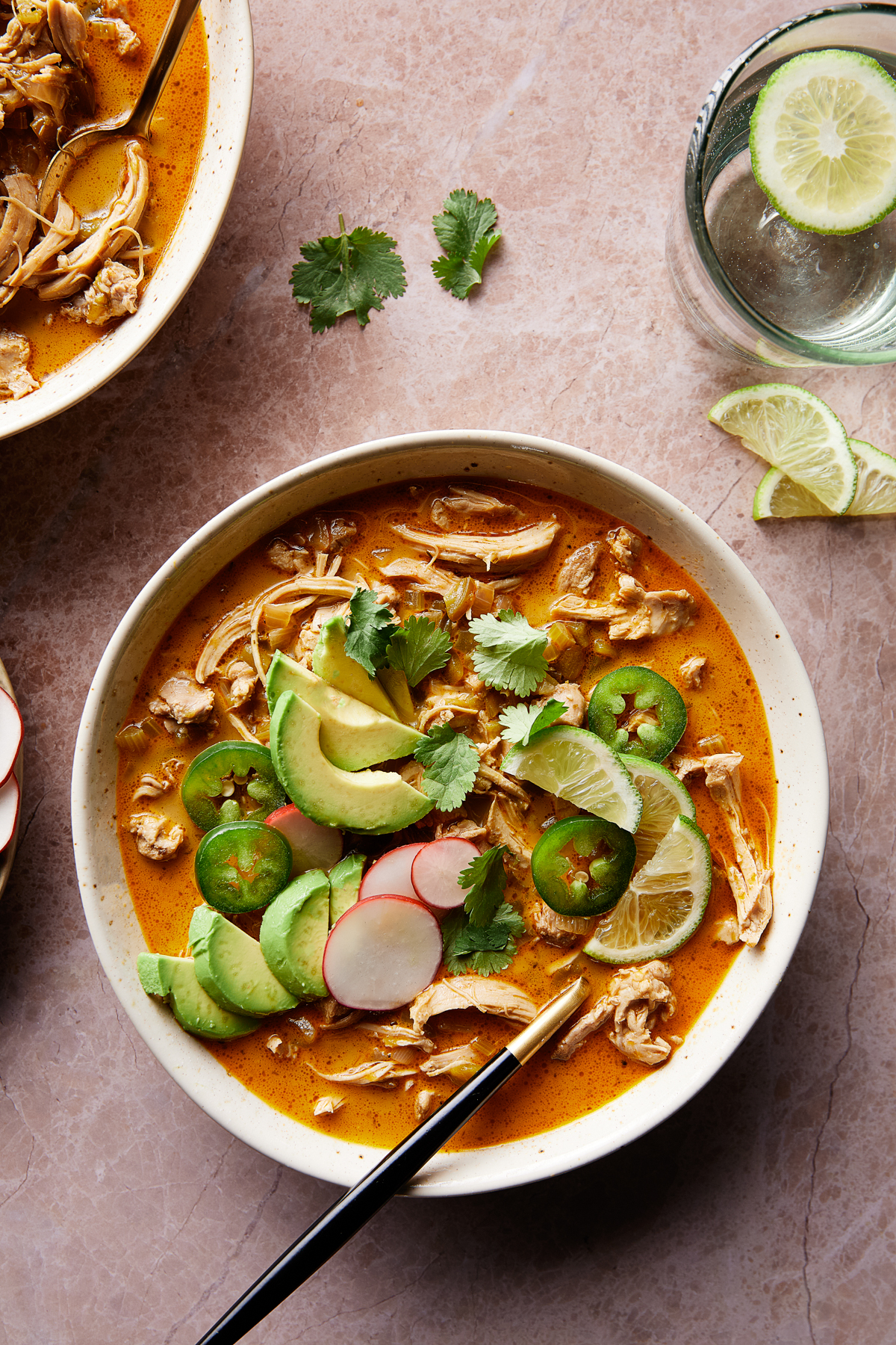 A bowl of spicy soup with shredded chicken, avocado slices, jalapeños, cilantro, radish, and lime. Next to it is a glass of water with lime slices. Everything is set on a textured pinkish surface.