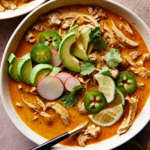 A bowl of keto white chicken chili with shredded chicken, avocado slices, jalapeños, cilantro, radish, and lime. Next to it is a glass of water with lime slices. Everything is set on a textured pinkish surface.
