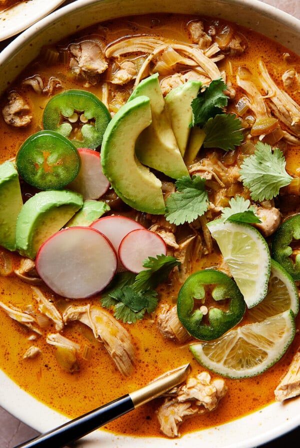 A bowl of keto white chicken chili with shredded chicken, avocado slices, jalapeños, cilantro, radish, and lime. Next to it is a glass of water with lime slices. Everything is set on a textured pinkish surface.