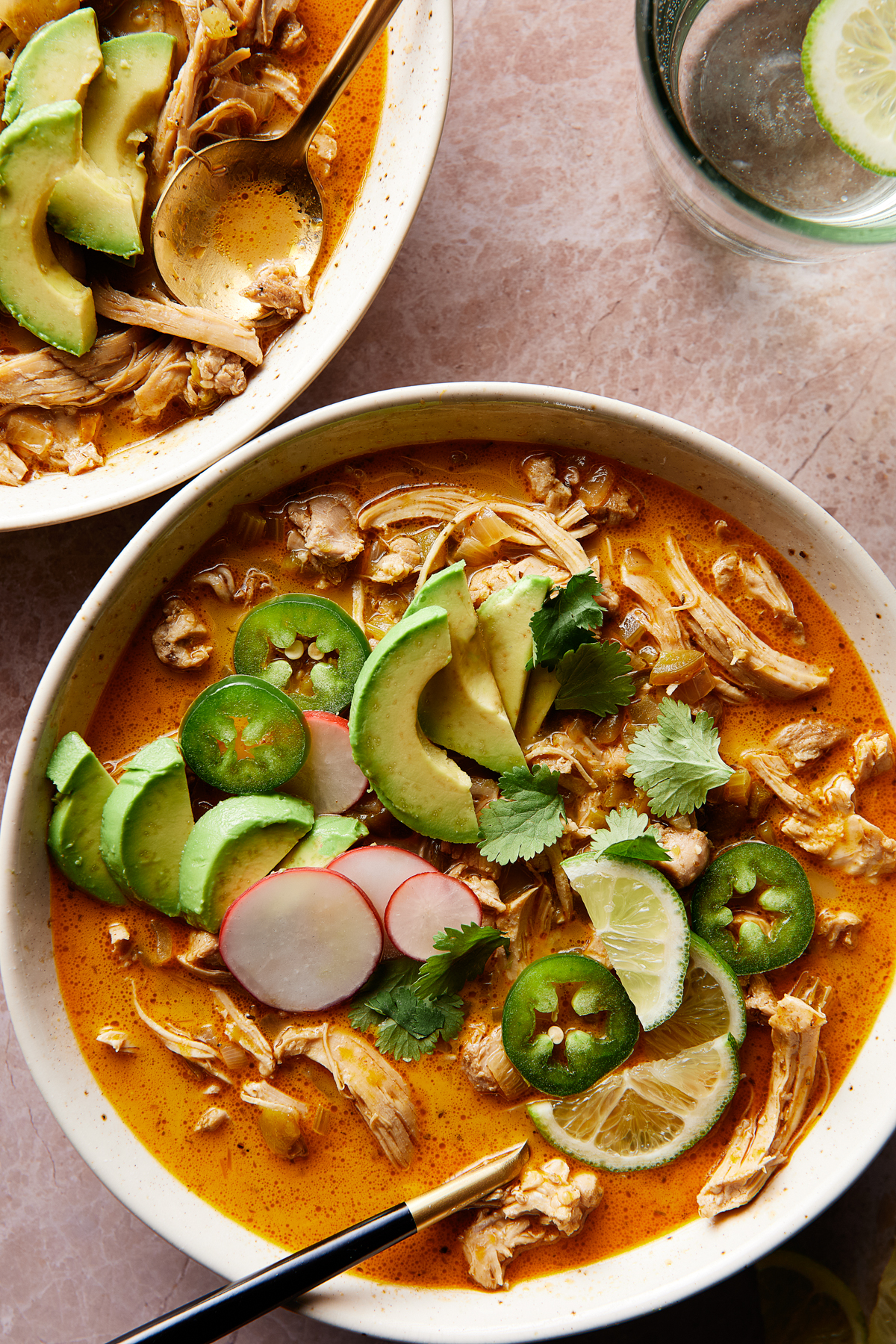 Two bowls of vibrant white chicken chili soup topped with sliced avocado, jalapeños, radishes, cilantro, and lime wedges. A glass of water with lime is seen nearby.