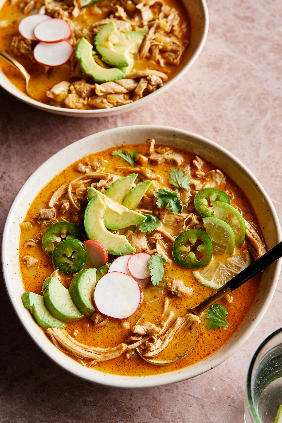 Two bowls of keto white chicken chili garnished with sliced avocado, jalapeños, radishes, cilantro, and lime. The soup has a rich, orange-tinted broth with shredded chicken, set on a marble countertop.
