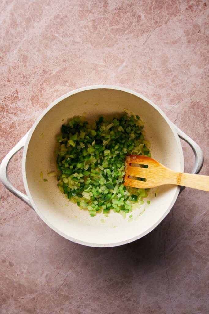 A white pot on a textured countertop contains finely chopped green bell peppers and onions being stirred with a wooden spatula.