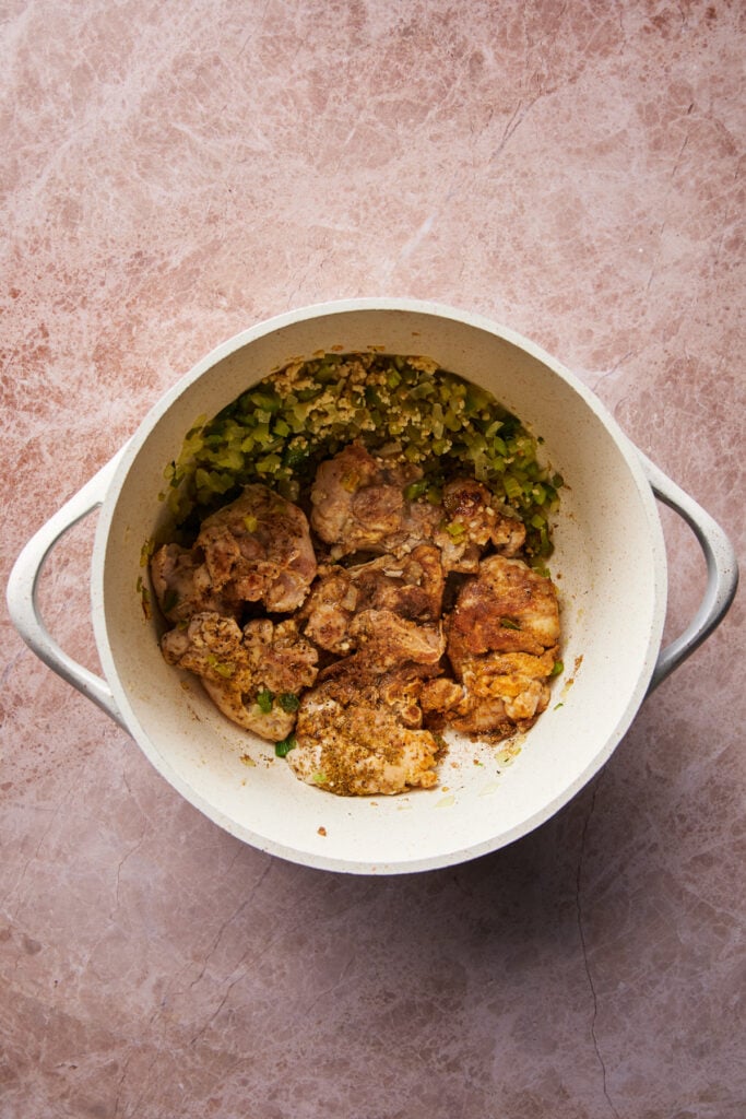A cooking pot containing seasoned pieces of chicken and chopped vegetables on a textured pink surface. The chicken pieces are partially cooked and surrounded by what appears to be onions and green peppers.