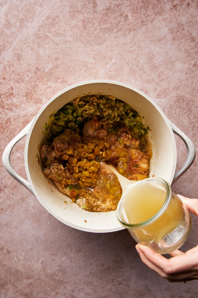 A hand pours broth from a glass jar into a pot containing seasoned meat, chopped green peppers, and spices. The pot is on a marbled countertop.