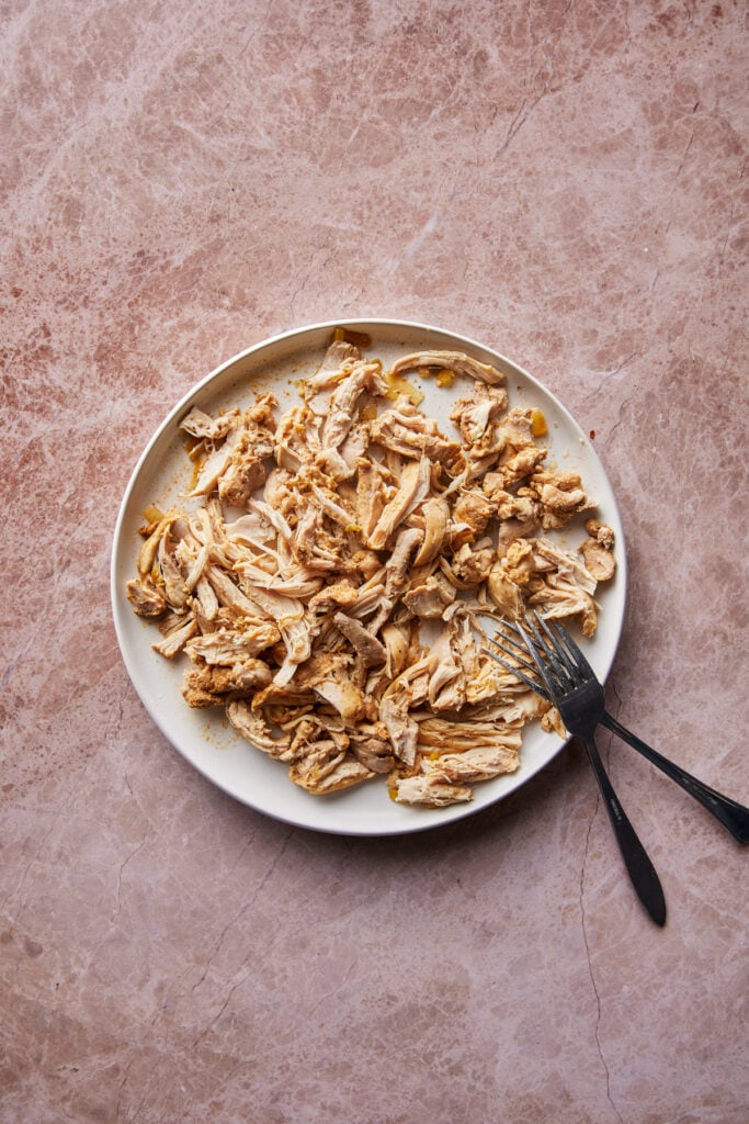 A plate of shredded cooked chicken on a marbled surface. A fork rests on the plate beside the chicken. The chicken appears seasoned and is spread out evenly across the white plate.