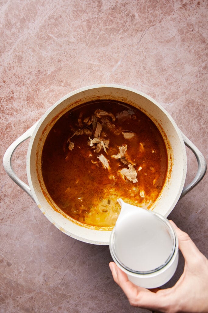 A white pot containing a reddish broth with pieces of chicken. A hand is pouring liquid from a glass measuring cup into the pot. The surface is a textured, light brown countertop.