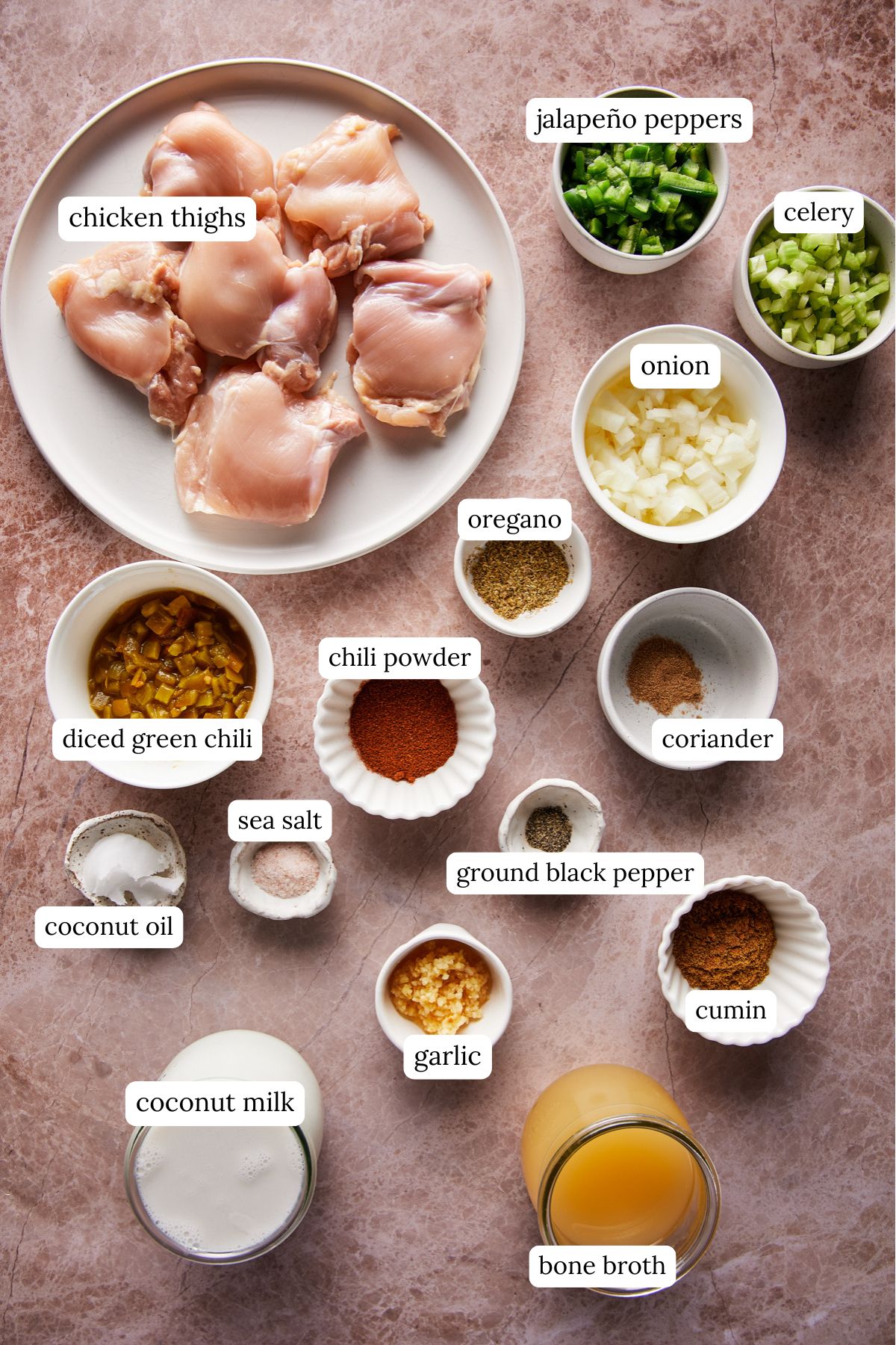 Top view of a kitchen counter with ingredients for a white chicken chili recipe: raw chicken thighs on a plate, small bowls of diced jalapeño peppers, celery, onion, oregano, ground coriander, chili powder, sea salt, ground black pepper, cumin, diced green chili, coconut oil, minced garlic, coconut milk, and a glass of bone broth.