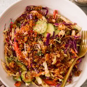 A vibrant crispy rice salad in a white bowl featuring shredded red and green cabbage, cucumber slices, red bell pepper, and crispy rice crumbles, topped with sesame seeds. A gold fork lies on the side. A jar of sesame seeds and a cut limes are nearby.