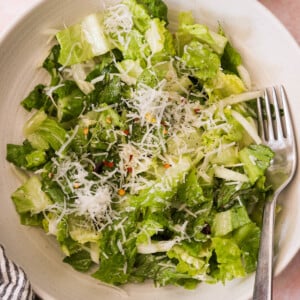 A bowl of Caesar salad with crisp romaine lettuce, grated cheese, pepper, and dressing. A fork rests along the edge of the white bowl.