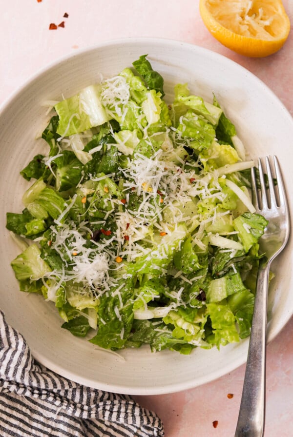 A bowl of Caesar salad with crisp romaine lettuce, grated cheese, pepper, and dressing. A fork rests along the edge of the white bowl.