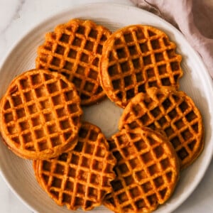 A white plate with six round, golden-brown sweet potato waffles arranged in a circular pattern on a marble surface. A soft, light purple cloth is draped to the right of the plate.