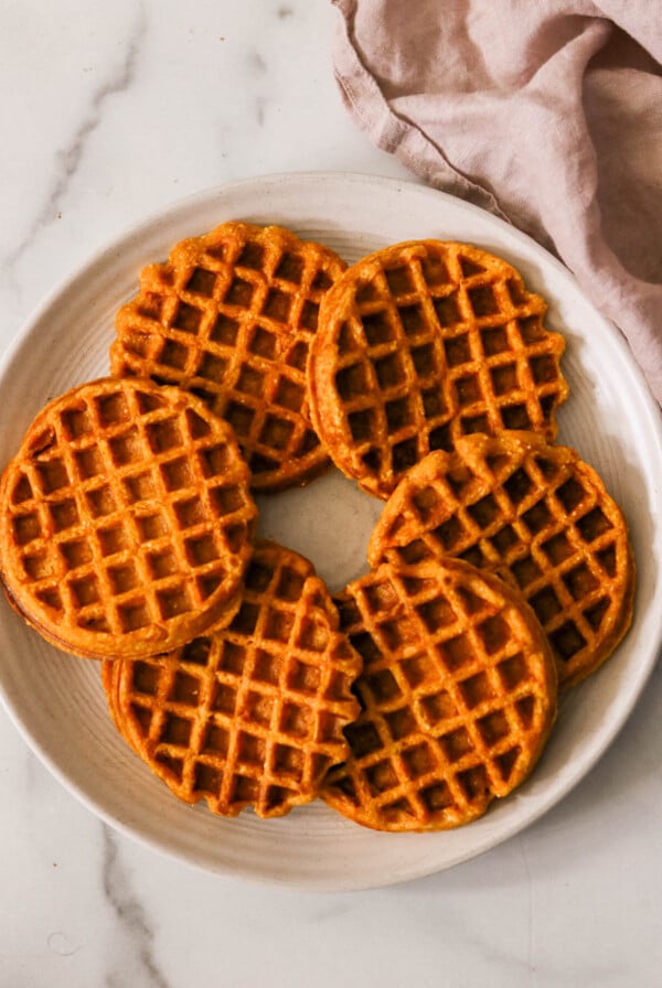 A white plate with six round, golden-brown sweet potato waffles arranged in a circular pattern on a marble surface. A soft, light purple cloth is draped to the right of the plate.