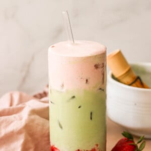 A tall glass filled with a layered drink featuring a pink foam on top, a green matcha middle with ice, and red strawberry puree at the bottom. A glass straw is inserted into the drink. Fresh strawberries and a bowl are in the background.