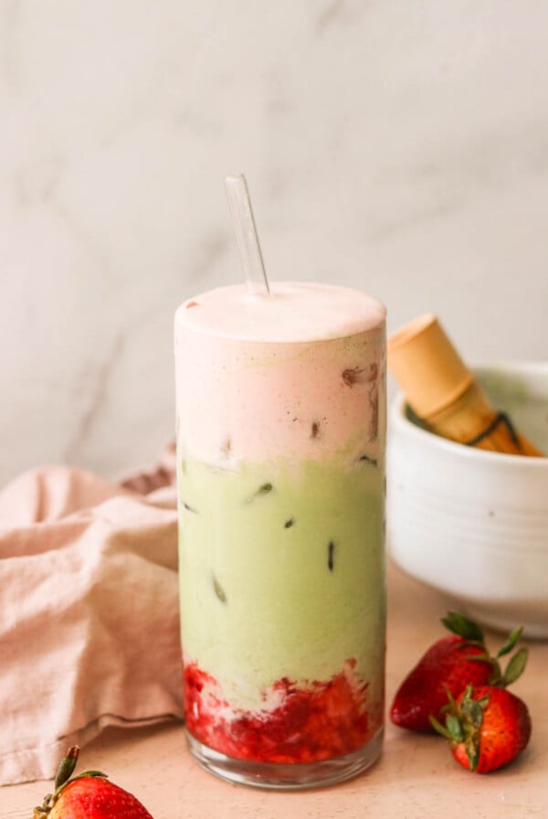 A tall glass filled with a layered drink featuring a pink foam on top, a green matcha middle with ice, and red strawberry puree at the bottom. A glass straw is inserted into the drink. Fresh strawberries and a bowl are in the background.