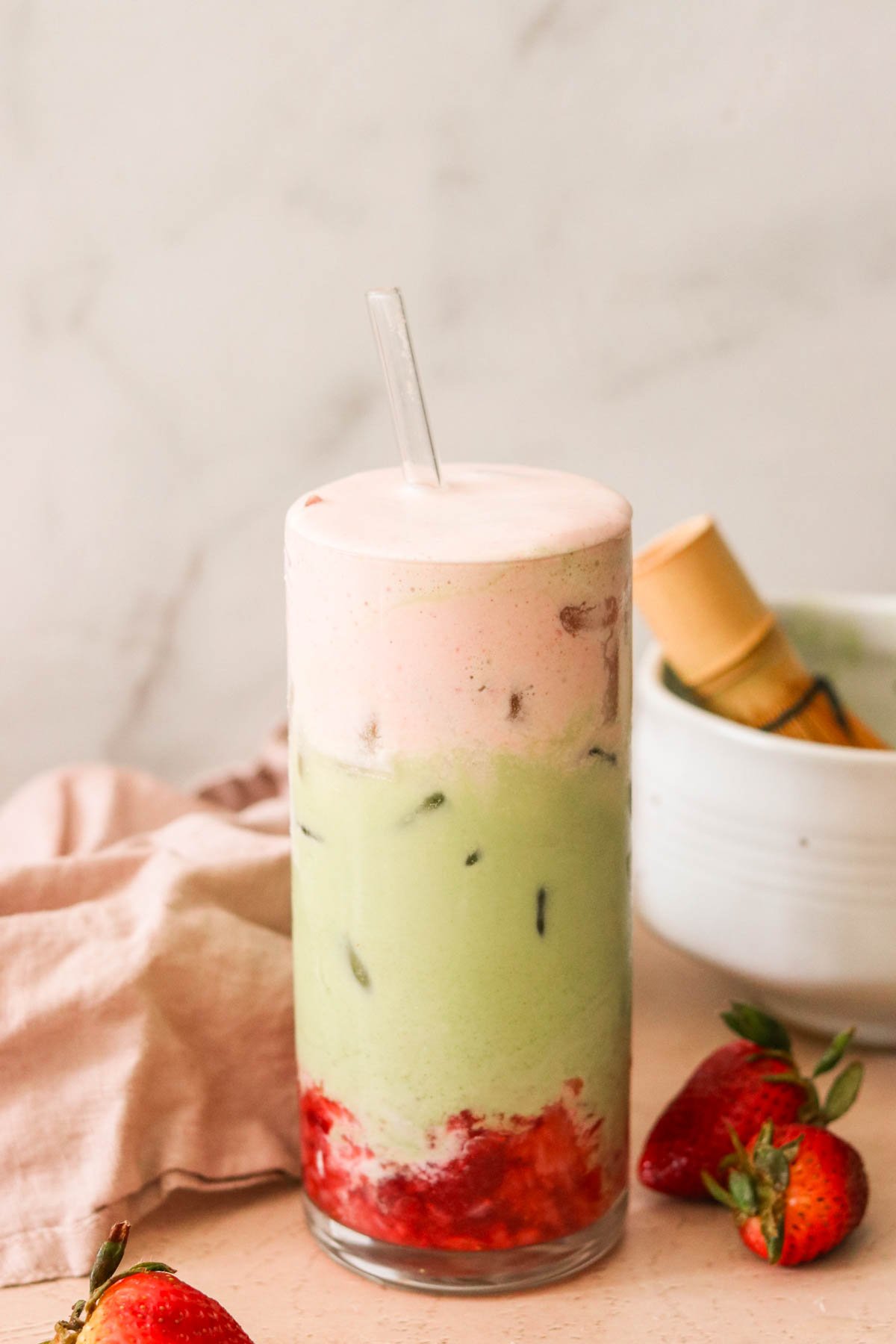 A tall glass filled with a layered drink featuring a pink foam on top, a green matcha middle with ice, and red strawberry puree at the bottom. A glass straw is inserted into the drink. Fresh strawberries and a bowl are in the background.