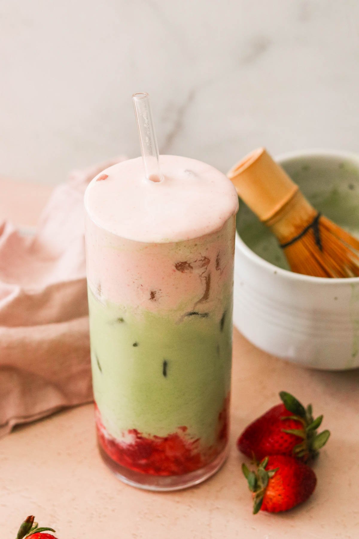 an iced strawberry match latte in a glass with ice next to a bowl with a bamboo matcha whisk inside. 