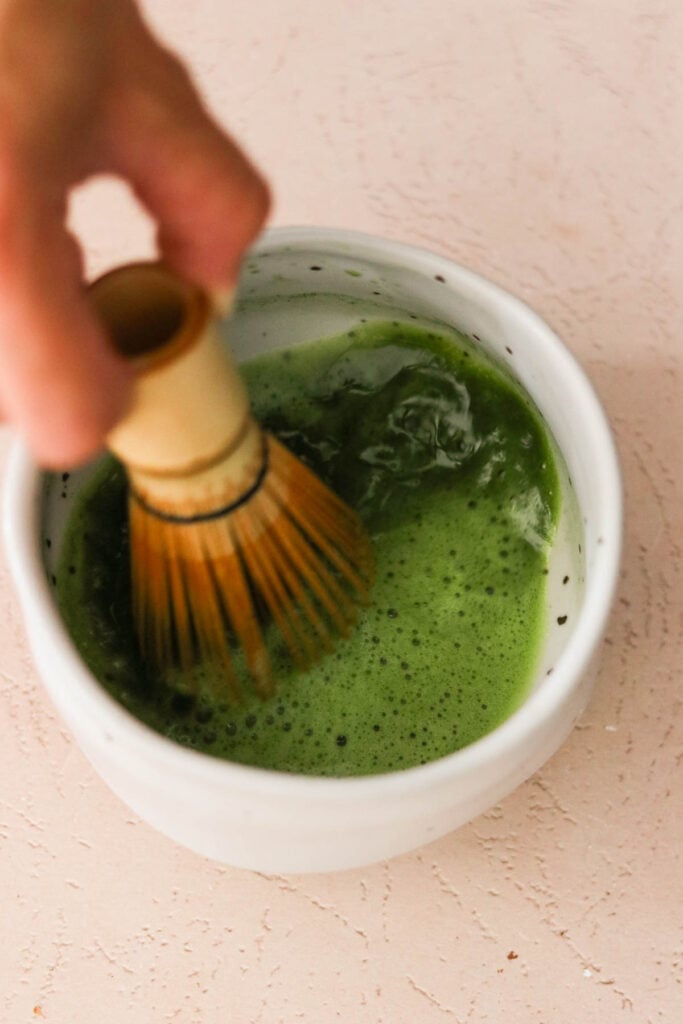 a bamboo whisk mixing matcha into hot water in a small bowl 